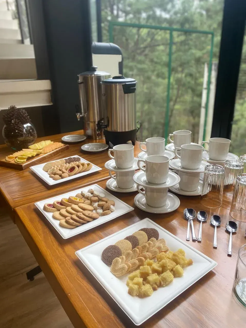 Té y pasteles dispuestos sobre una mesa de madera junto a una ventana, con tazas, cucharas y cafeteras.
