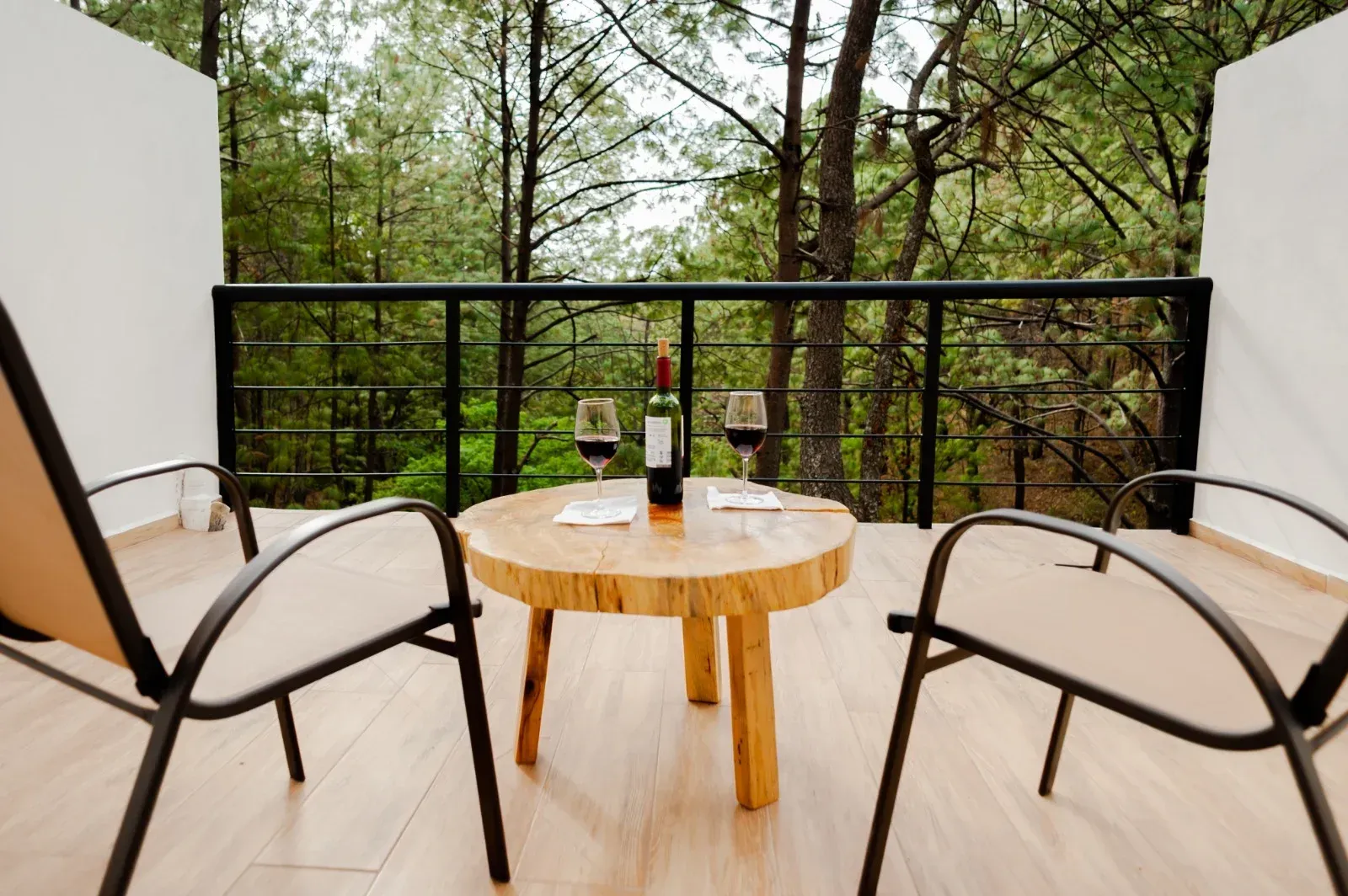 Zona de estar en el balcón con dos sillas, mesa redonda de madera y botellas de vino con vistas a los árboles.