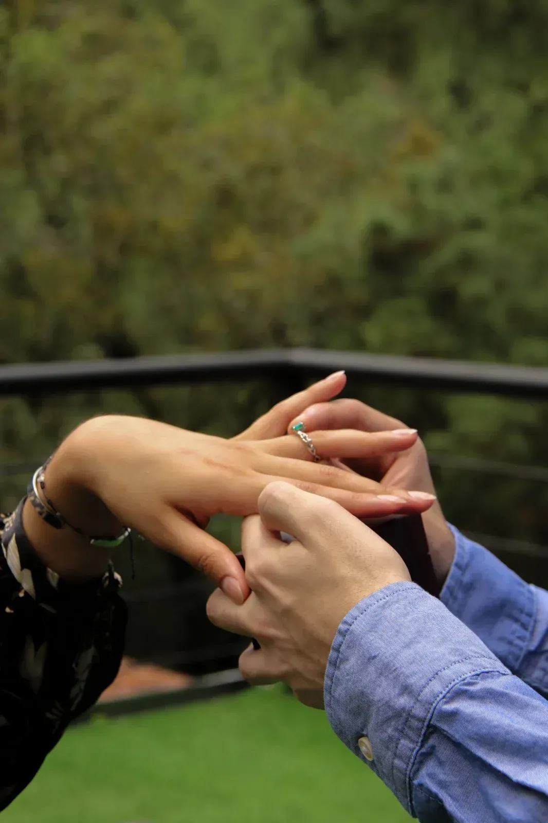Dos personas tomadas de la mano con delicadeza al aire libre, con un fondo verde borroso y una barandilla.