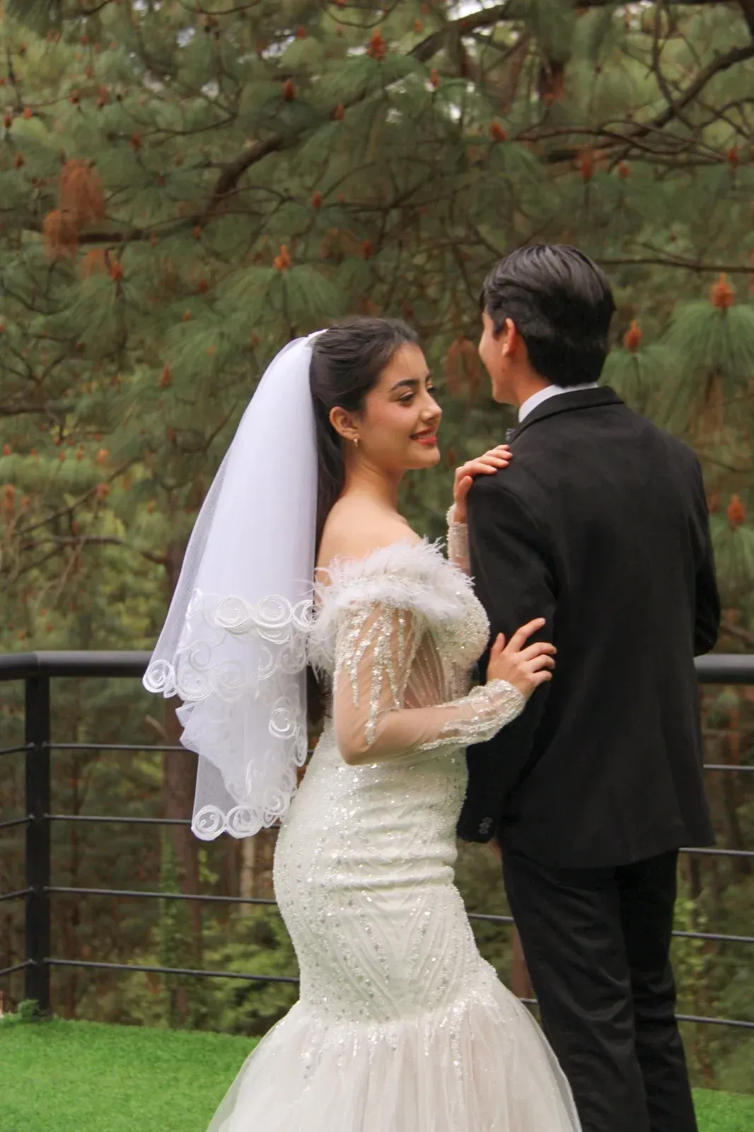 Novios abrazándose al aire libre junto a una valla de madera, con pinos al fondo.