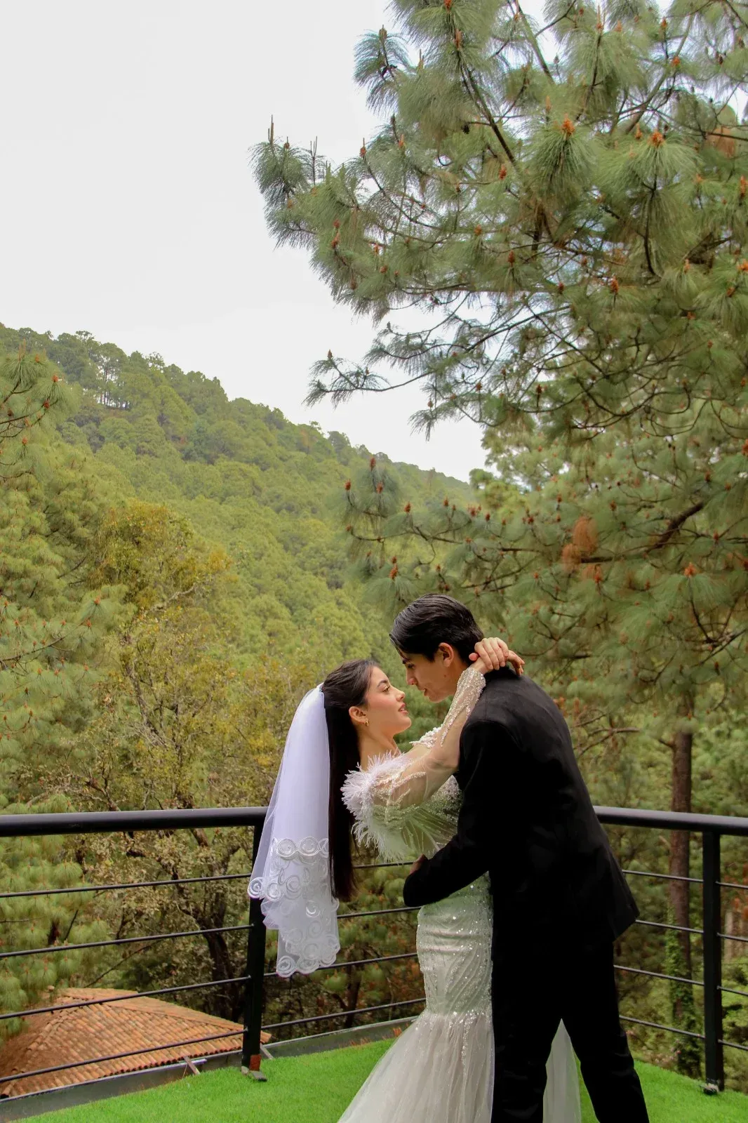Una pareja vestida de novios se abraza en un balcón panorámico con colinas boscosas de fondo.