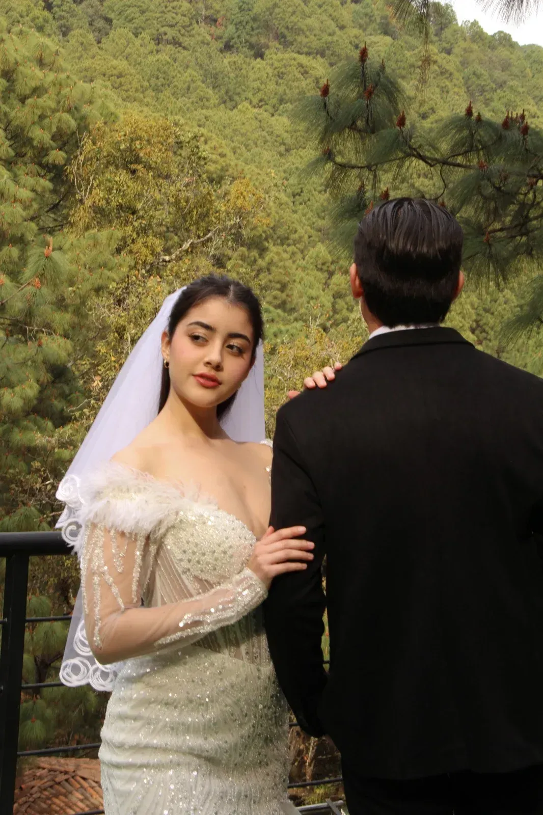 Novia vestida de blanco y velo, está de pie junto al novio, que lleva un traje negro, en una terraza en el bosque