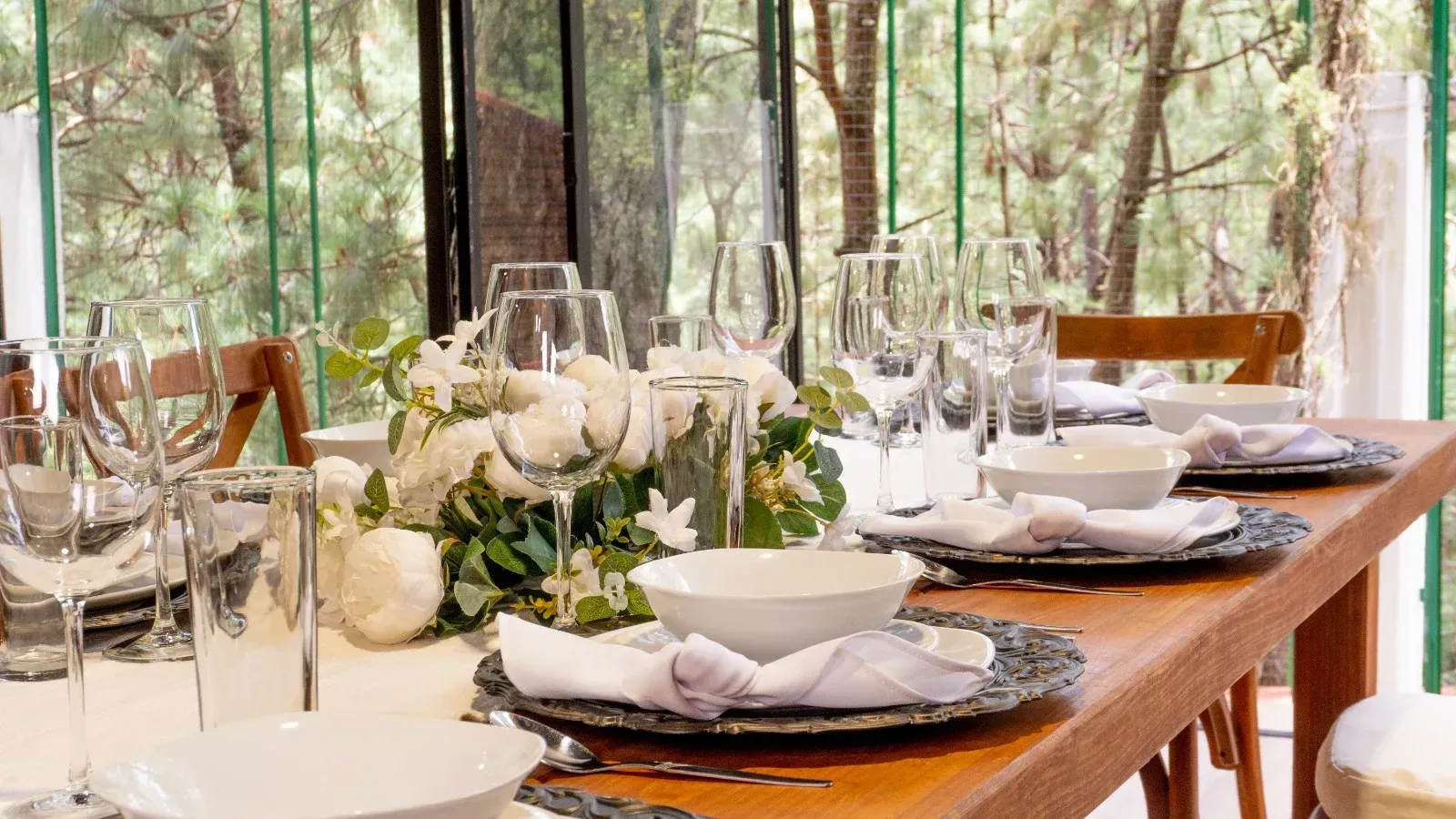 Elegante mesa de comedor con vajilla blanca, flores y cristalería en un luminoso entorno boscoso.