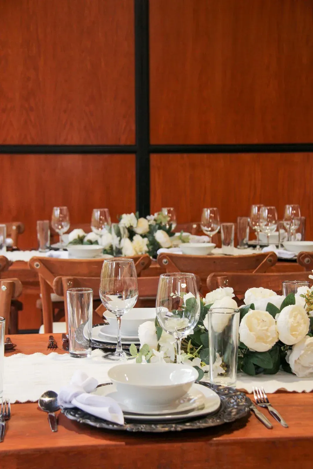 Elegante mesa de banquete puesta con centro de mesa floral, cristalería y platos blancos frente a cálida pared de madera