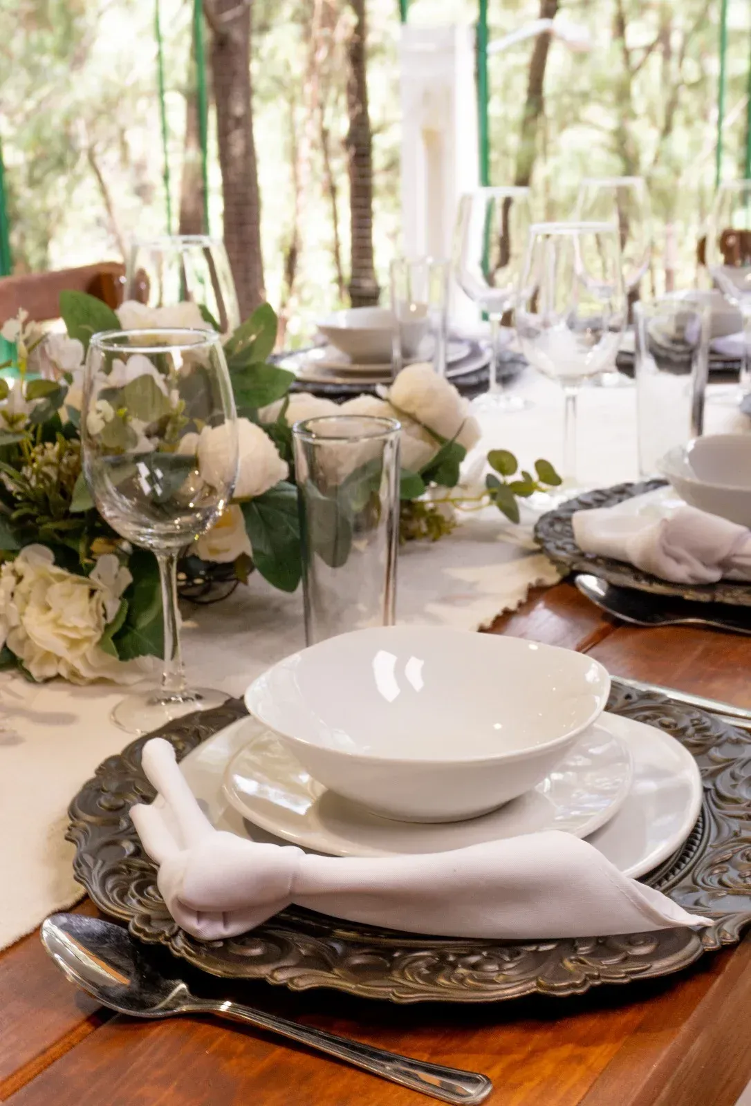 Elegante conjunto de mesa y sillas para comer al aire libre con vajilla blanca, vasos y arreglos florales en color crema.