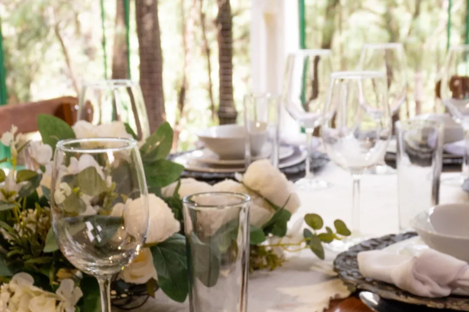 Elegante mesa de comedor al aire libre con copas de vino, flores blancas y vegetación en un entorno de jardín soleado.