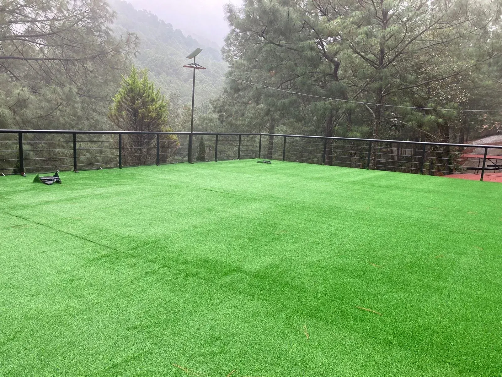 Patio de césped artificial con valla negra, con vistas a colinas boscosas envueltas en niebla.