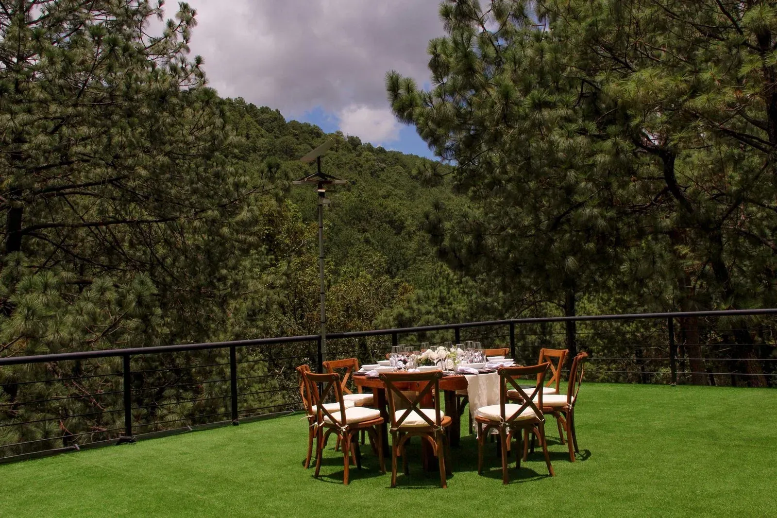 Patio exterior con mesa y sillas de madera sobre césped verde, rodeado de árboles y una barandilla negra.