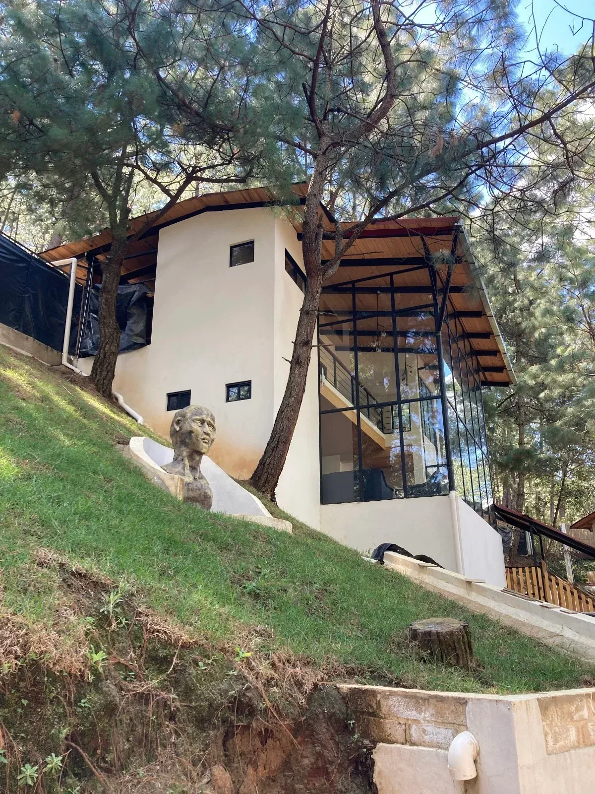 Pequeño edificio blanco con techo de cristal inclinado en una ladera cubierta de hierba entre árboles altos.