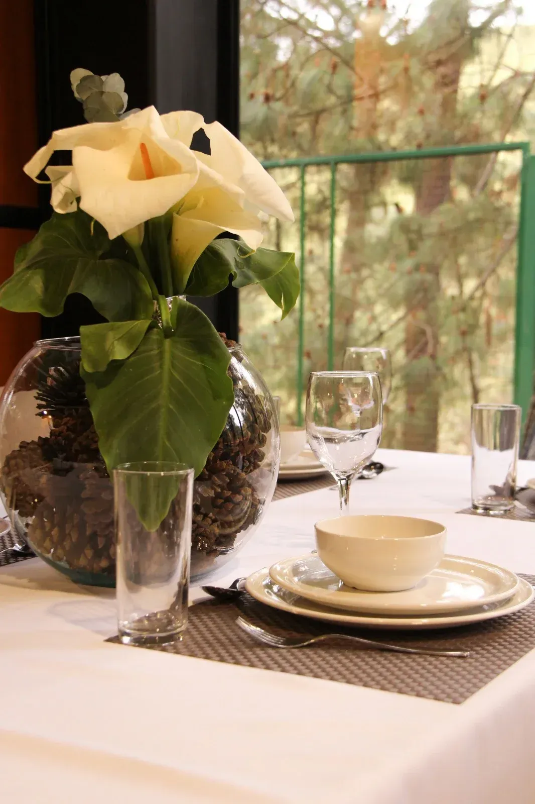 Elegante mesa puesta con flores blancas en un jarrón, copas de vino y platos apilados junto a una ventana.