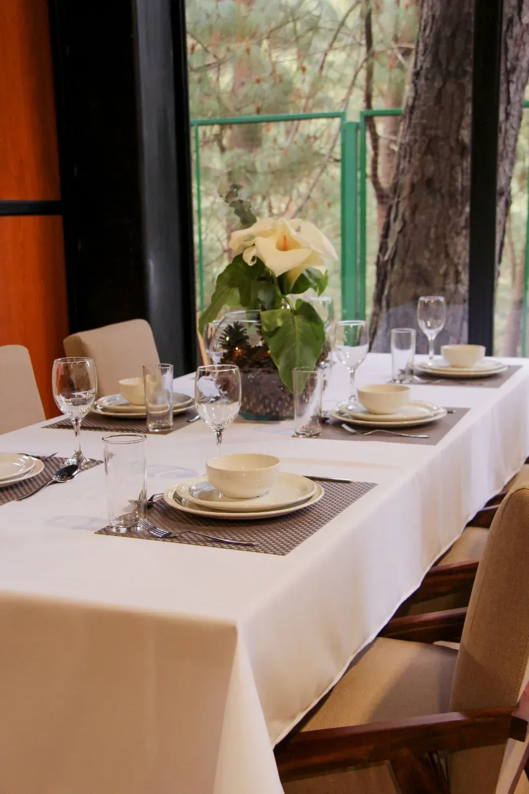 Elegante mesa de comedor puesta con manteles blancos, velas, cristalería y un centro de mesa floral junto a una ventana.