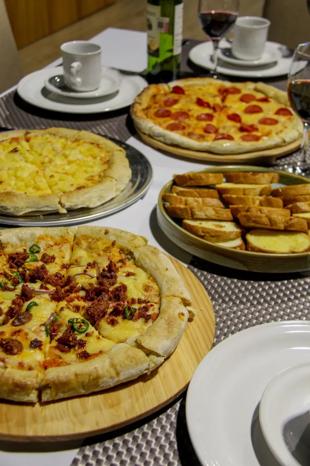 Varias pizzas y pan tostado sobre una mesa con platos, tazas y copas de vino.