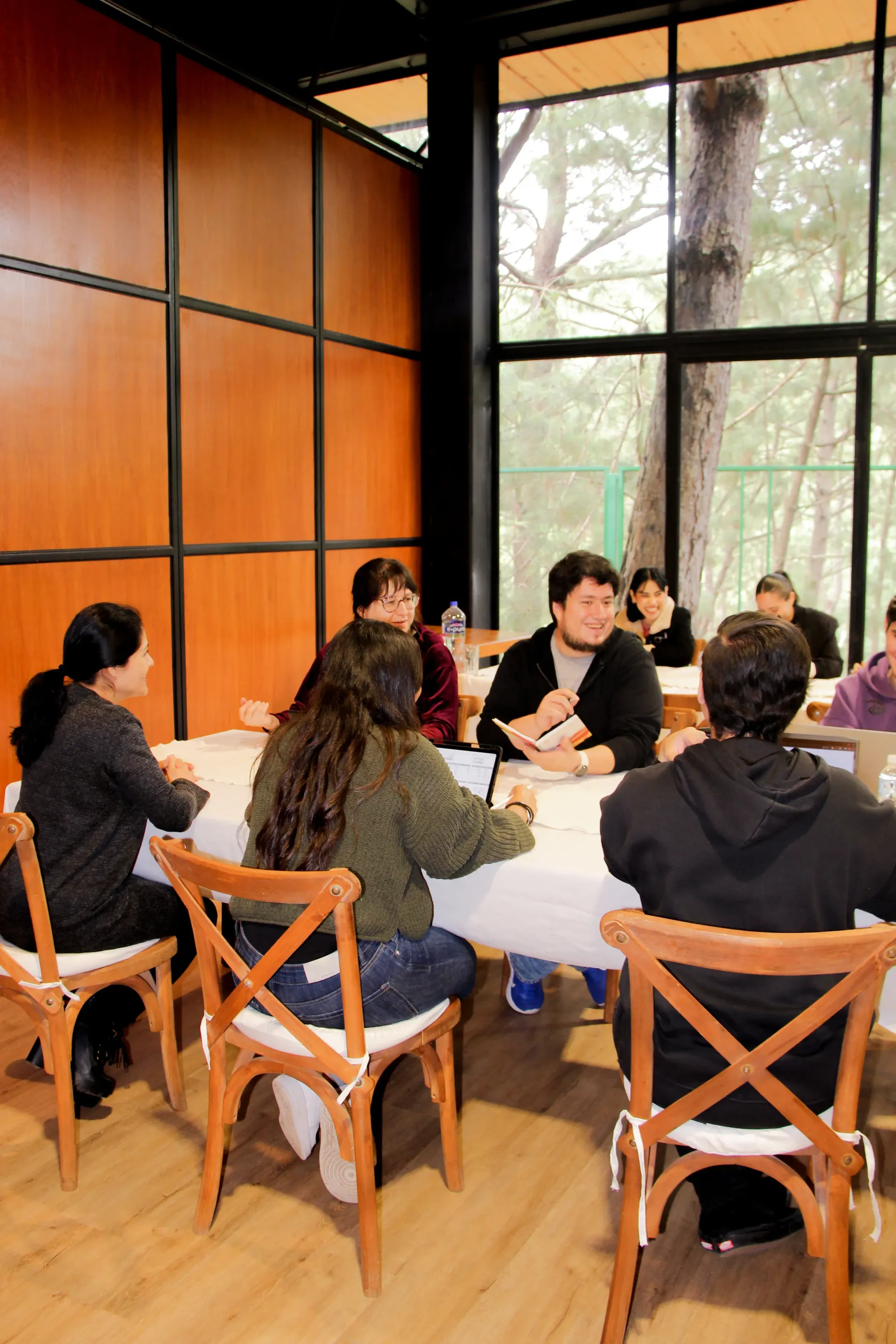 Personas sentadas alrededor de una mesa en un salón con grandes ventanales y paredes revestidas de paneles de madera