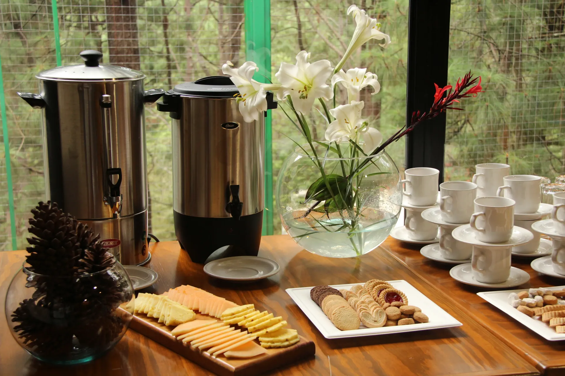 Mesa de buffet con cafeteras, tazas, pasteles y flores junto a una ventana.