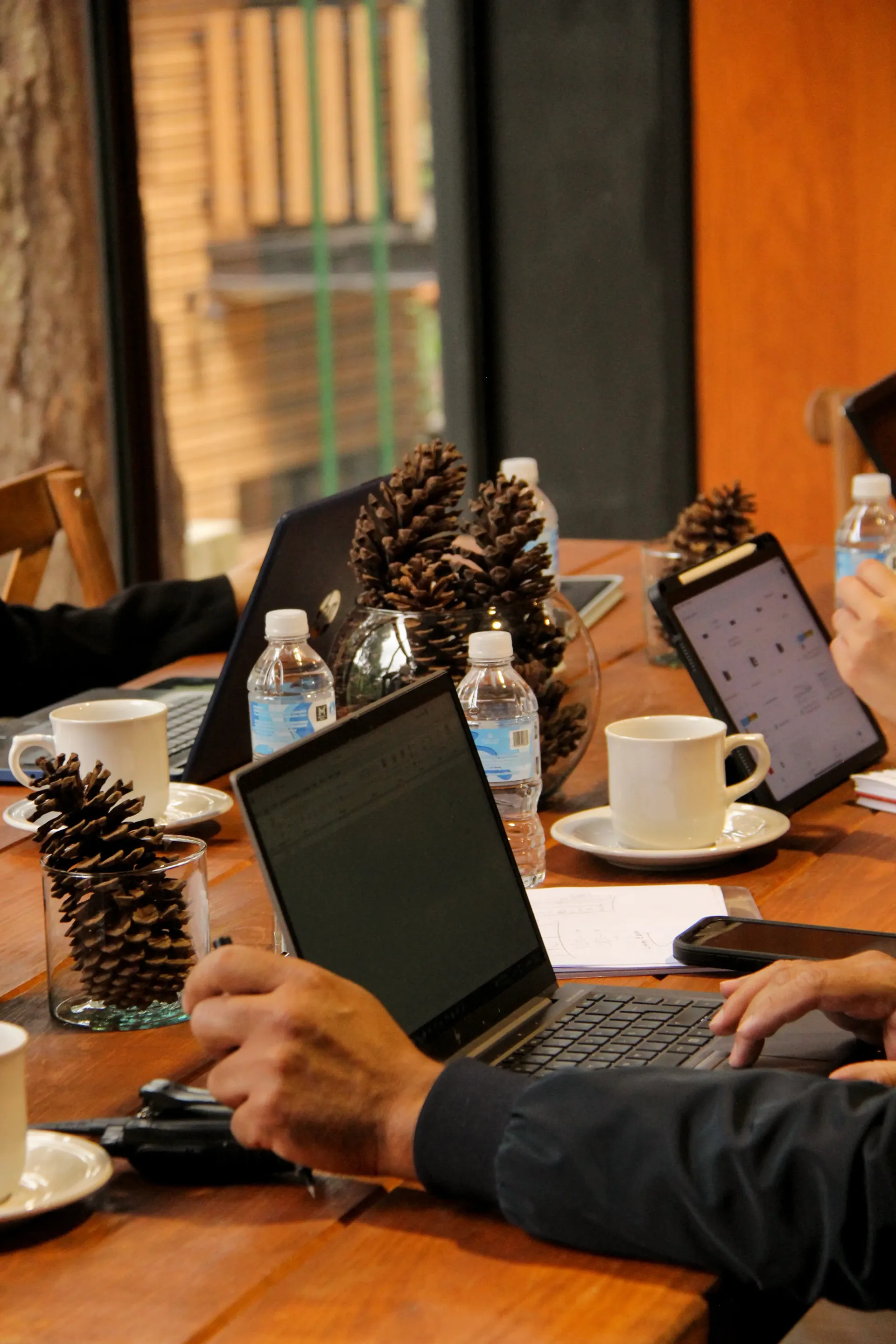 Personas trabajando en sus portátiles en una mesa de café de tonos cálidos, decorada con piñas y tazas de café.