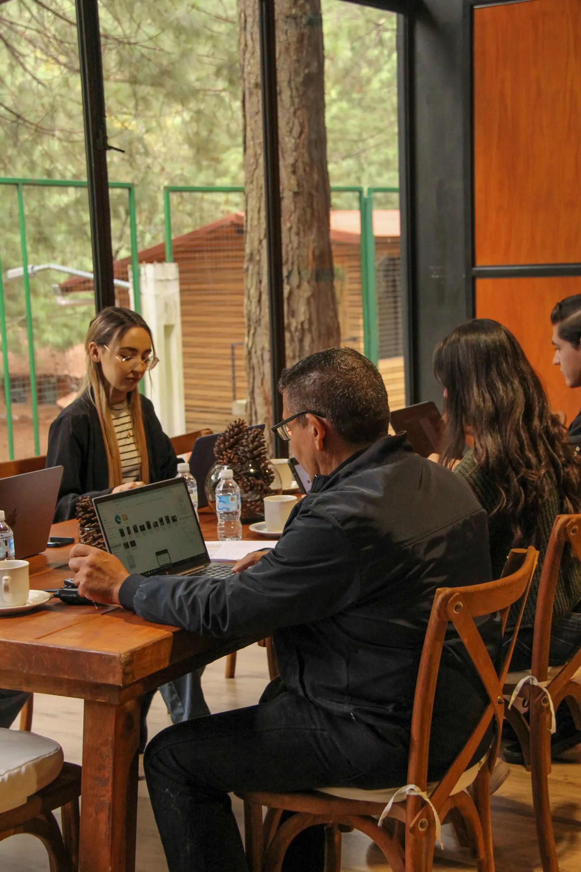 Personas sentadas en una mesa de madera en una cafetería con paredes de cristal tomando café y trabajando en laptops.