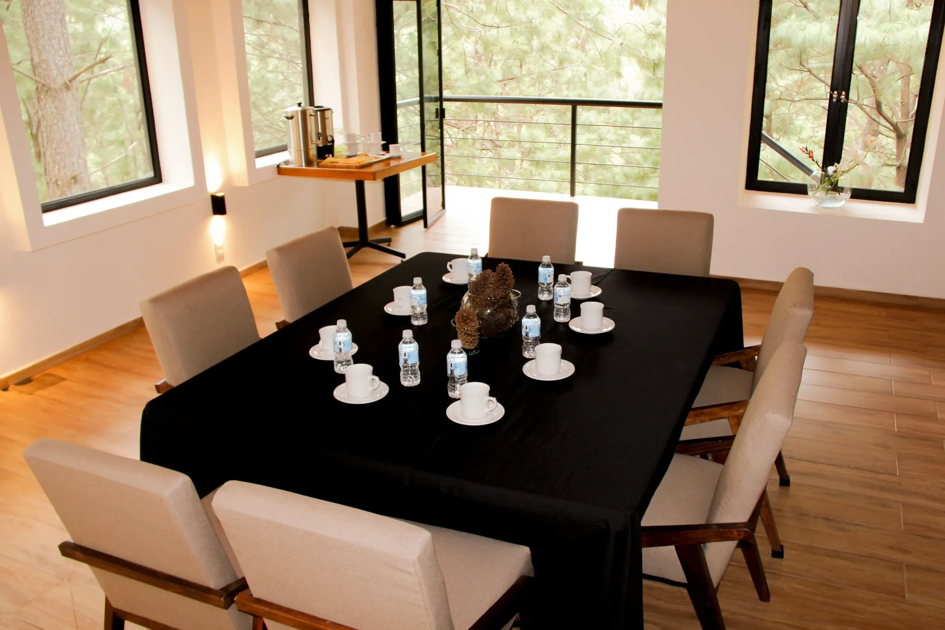 Comedor con una mesa cubierta con un mantel negro, preparada para tomar el té o el café, cerca de grandes ventanales.