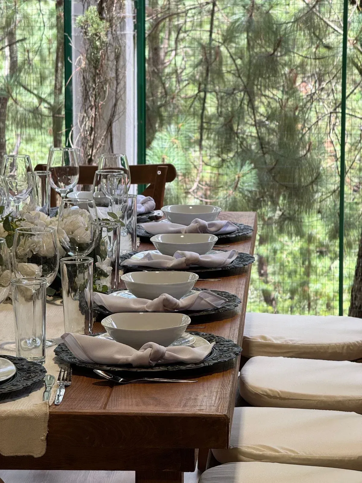 Prepara una mesa de comedor con vajilla y cristalería blancas junto a una ventana con vistas a los árboles.