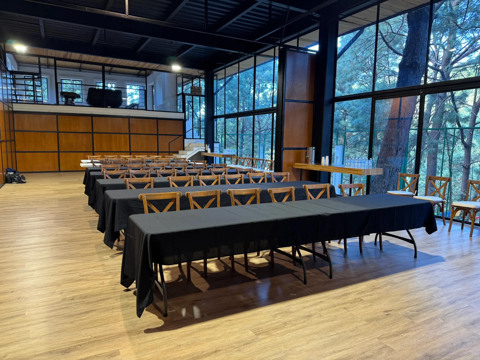 Salón de banquetes vacío con mesas largas, manteles negros y grandes ventanales con vistas a los árboles.