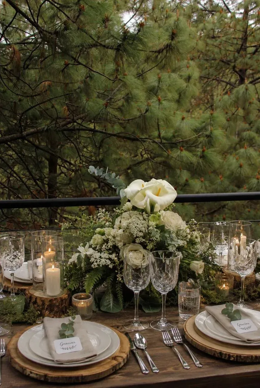 Elegante mesa de exterior con flores blancas, velas y platos con borde dorado sobre un fondo de pino.