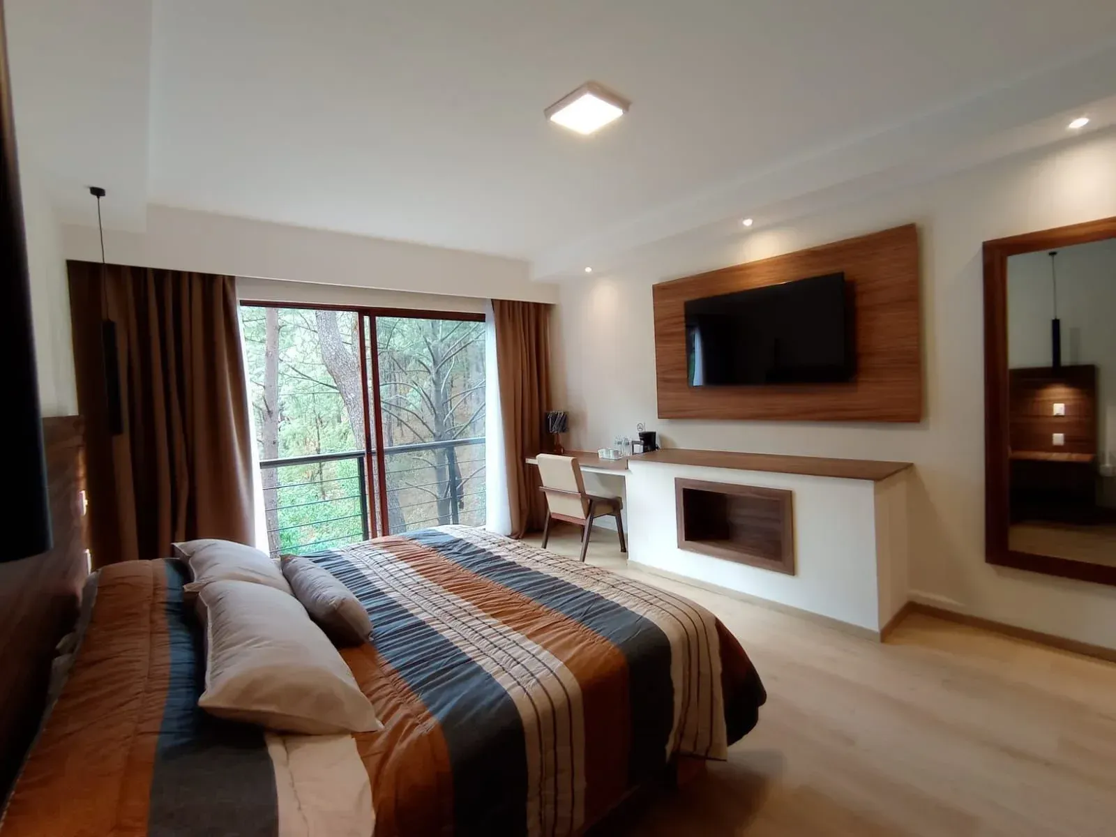 Dormitorio moderno con colcha a rayas, televisor en la pared, escritorio junto a la ventana y detalles en madera cálida