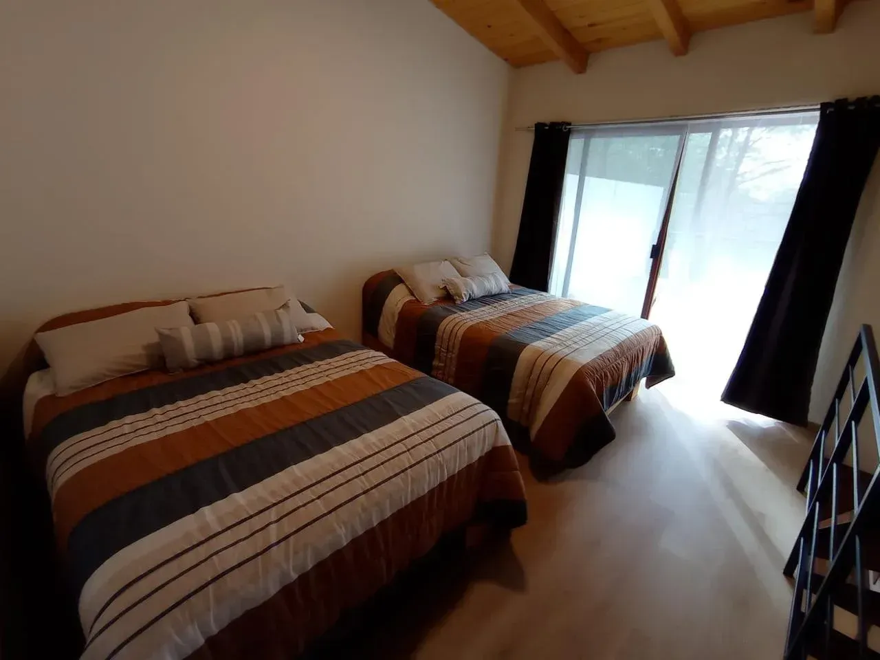 Habitación pequeña con dos camas individuales, mantas a rayas y puertas corredizas de cristal que dejan entrar mucha luz.