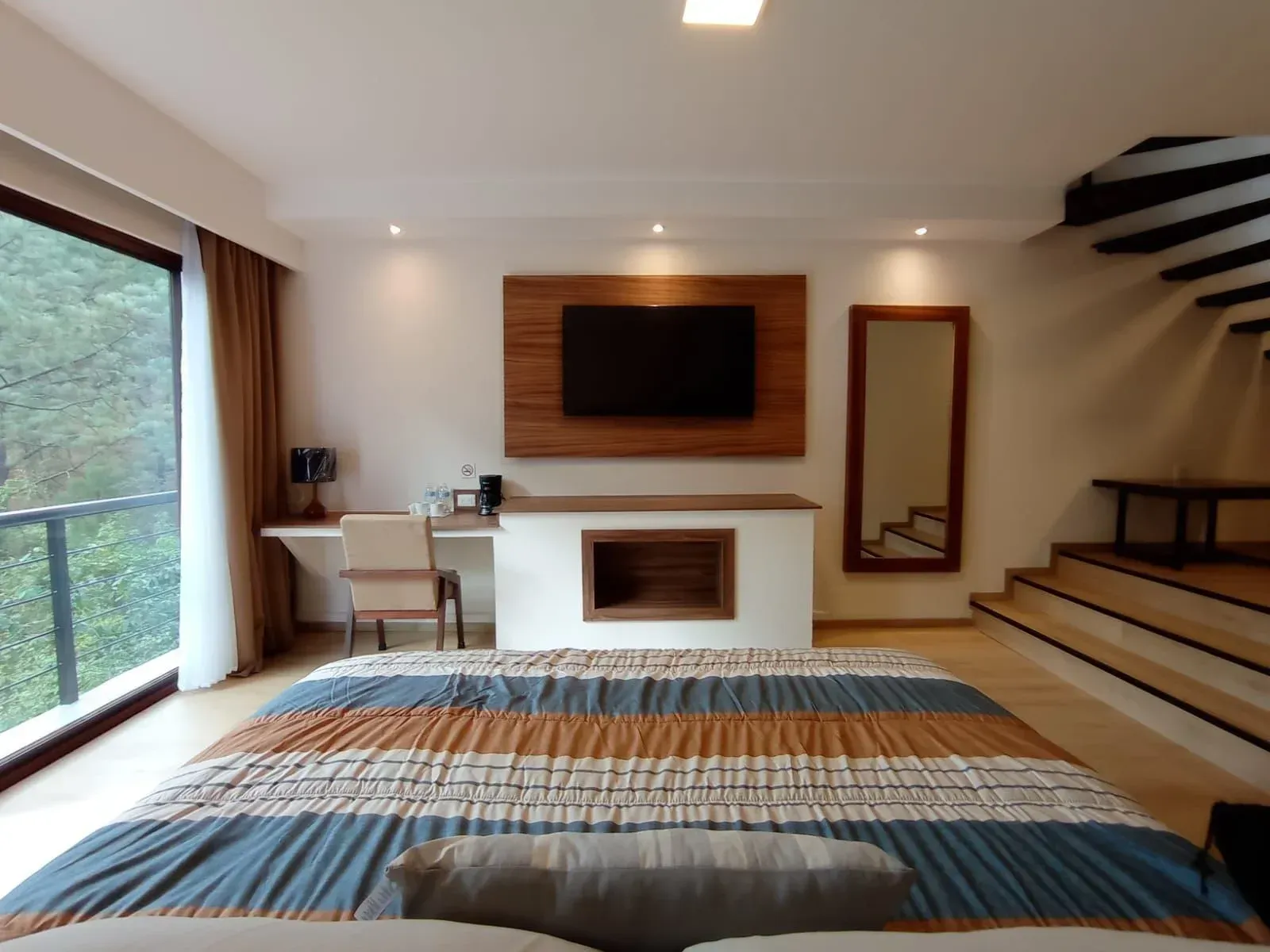 Dormitorio moderno con cama grande, t.v. en la pared, escritorio, espejo y escalera junto a una ventana con balcón.