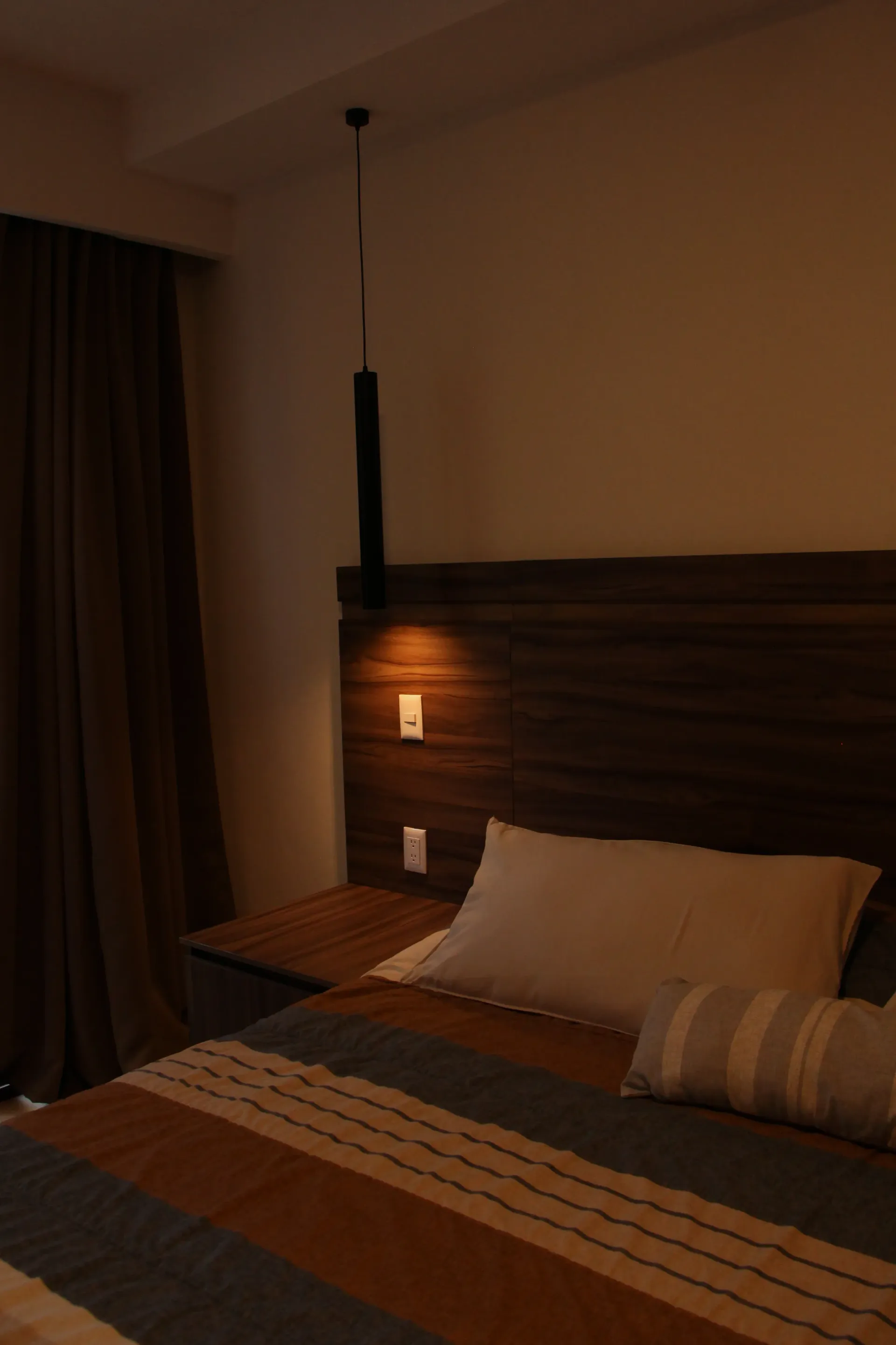 Habitación de hotel con iluminación tenue, cama, lámpara de noche y cortinas.