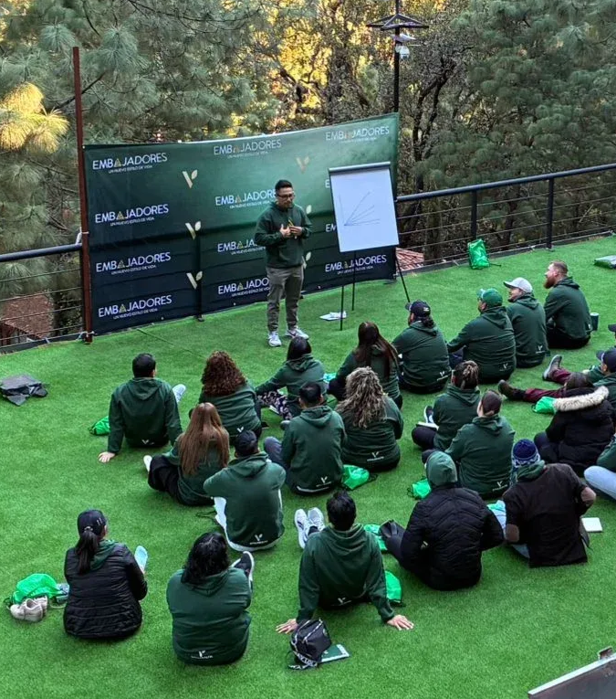 Presentación grupal al aire libre sobre césped, con un orador y público sentado frente a un telón de fondo y un rotafolio.