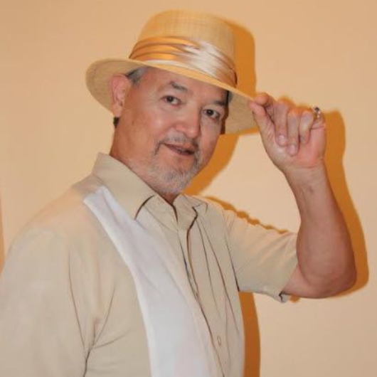 Man in light-colored hat and shirt, holding hat brim, smiling, against a beige background. Dan Mendoza