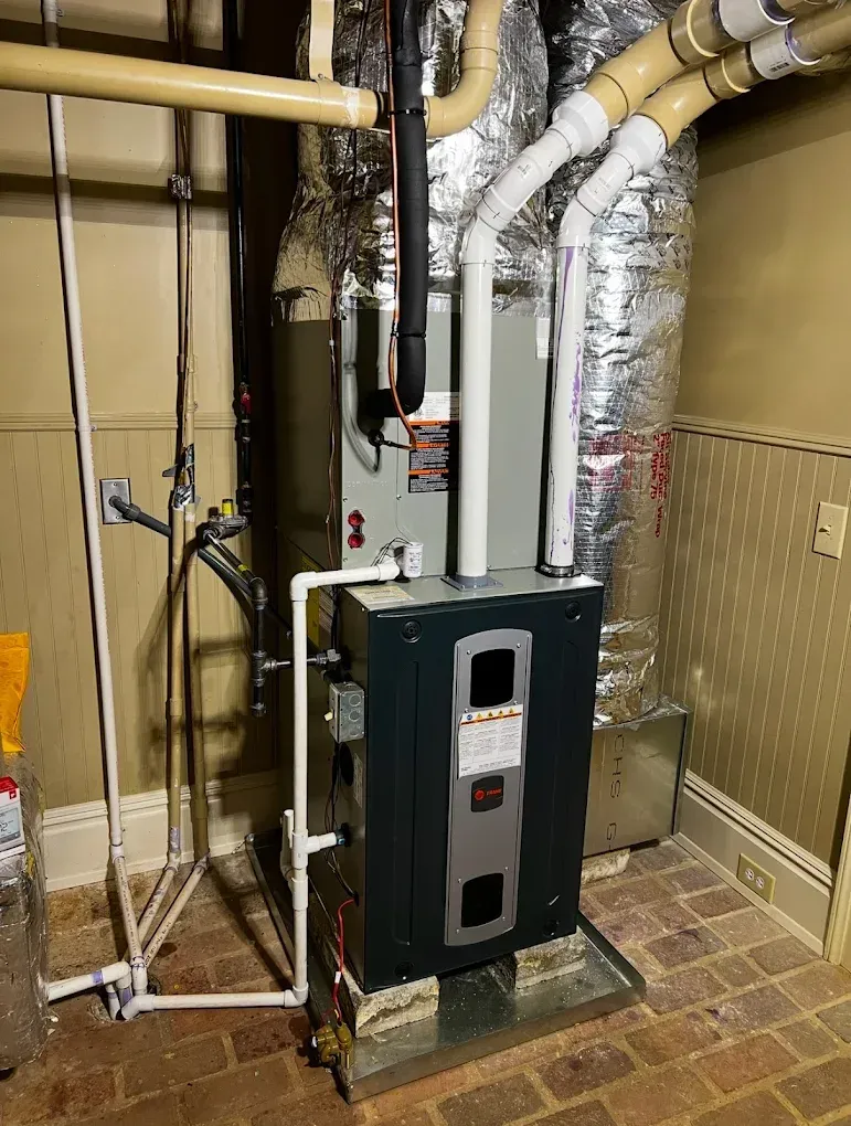Basement furnace with white ductwork, insulated pipes, and exposed utility lines against a beige wall.