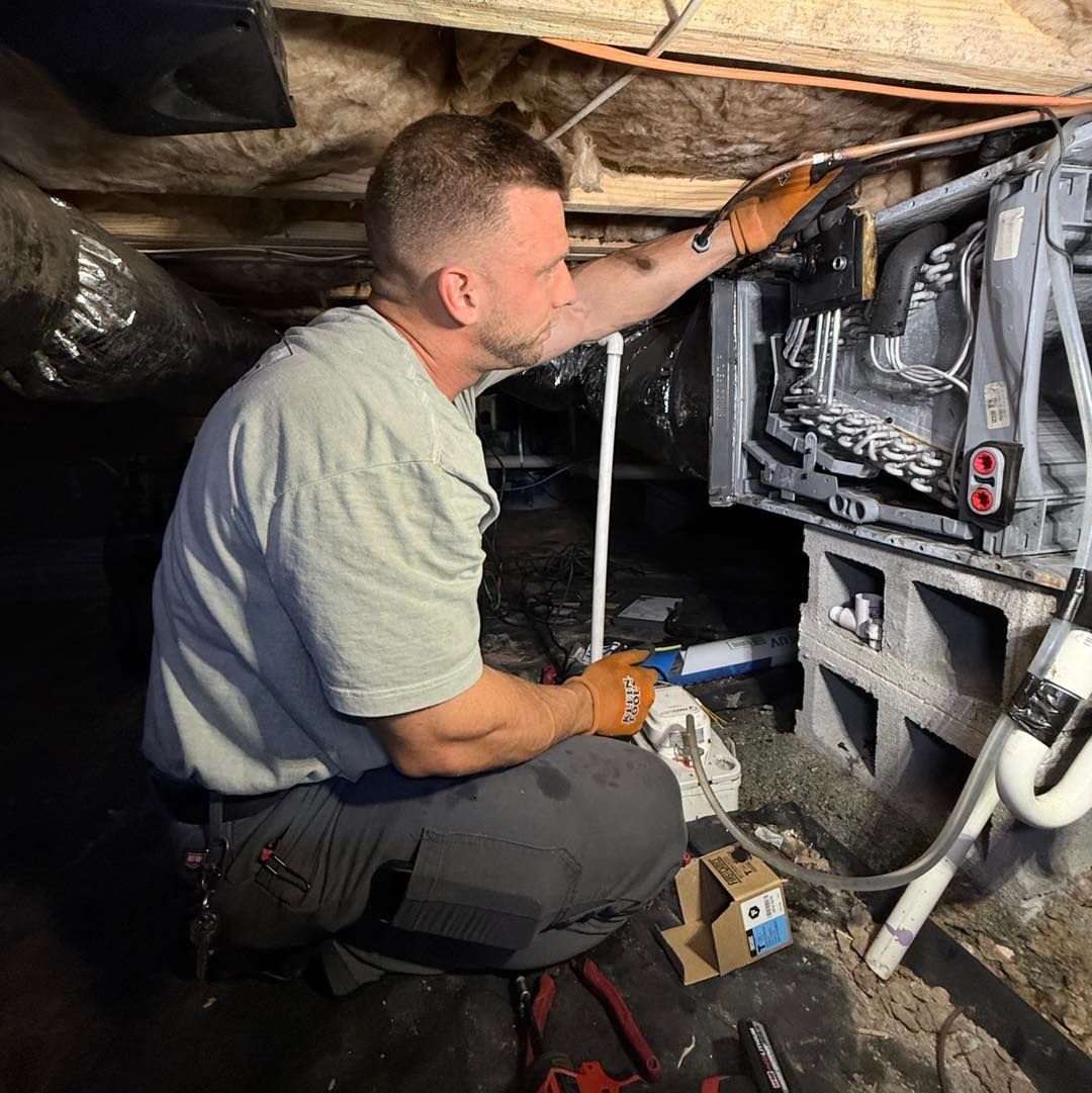 Technician inspecting HVAC unit in a crawlspace, kneeling beside exposed ducts and pipes.