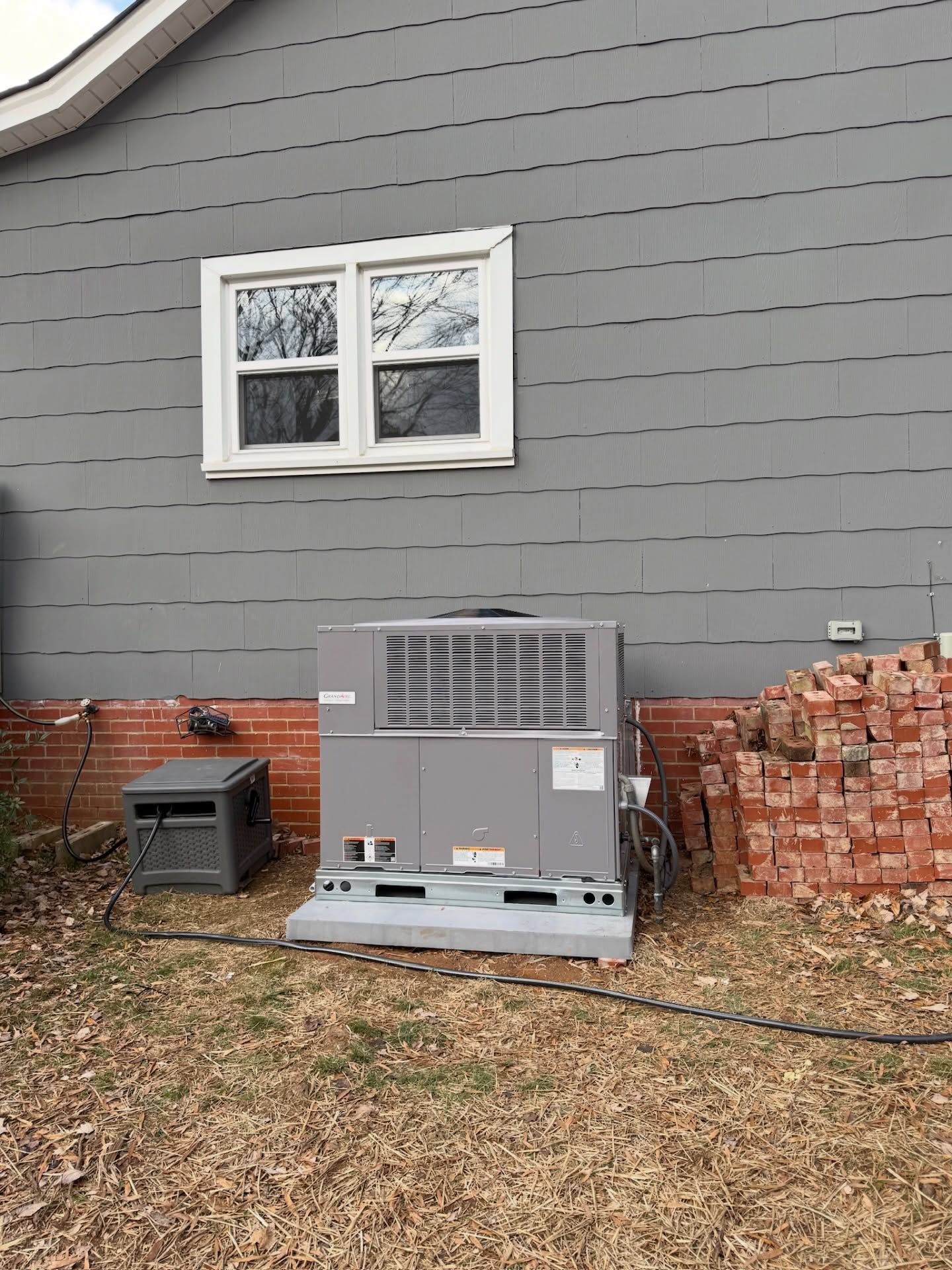 Gray house exterior with a large HVAC unit beside a stack of red bricks and a small black crate.