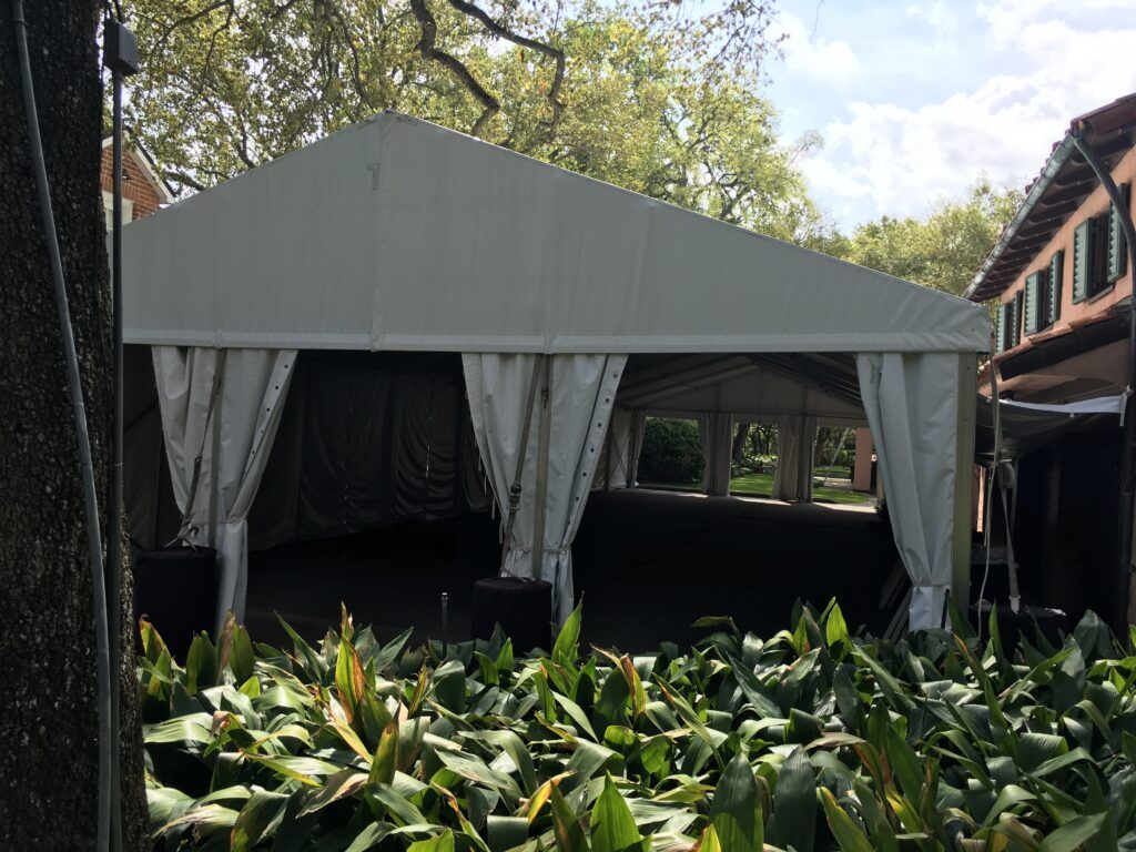 White tent in a garden setting, viewed from a bed of green plants. Buildings visible in the background.