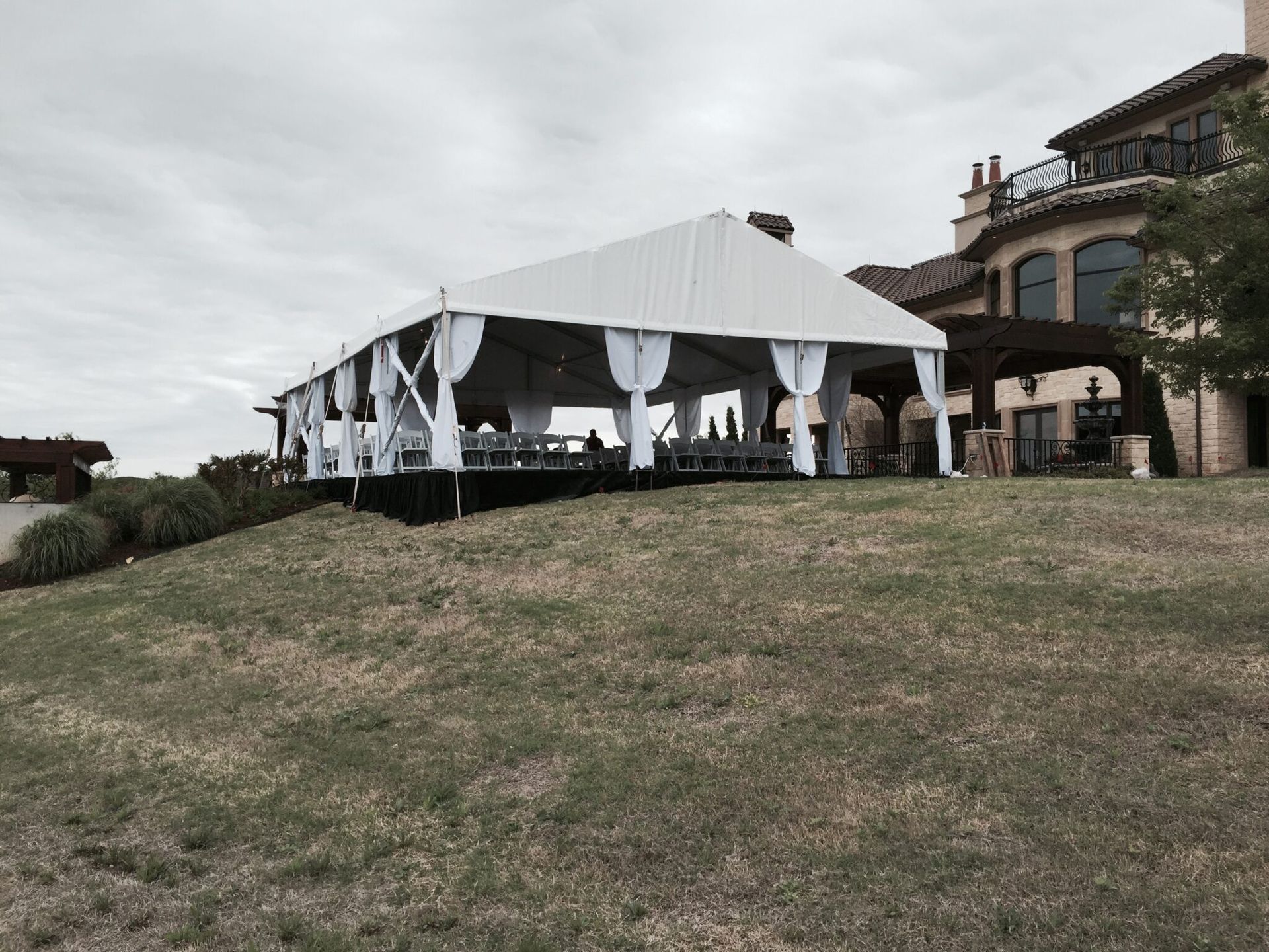 White tent set up on a grassy hill near a large house with a cloudy sky.