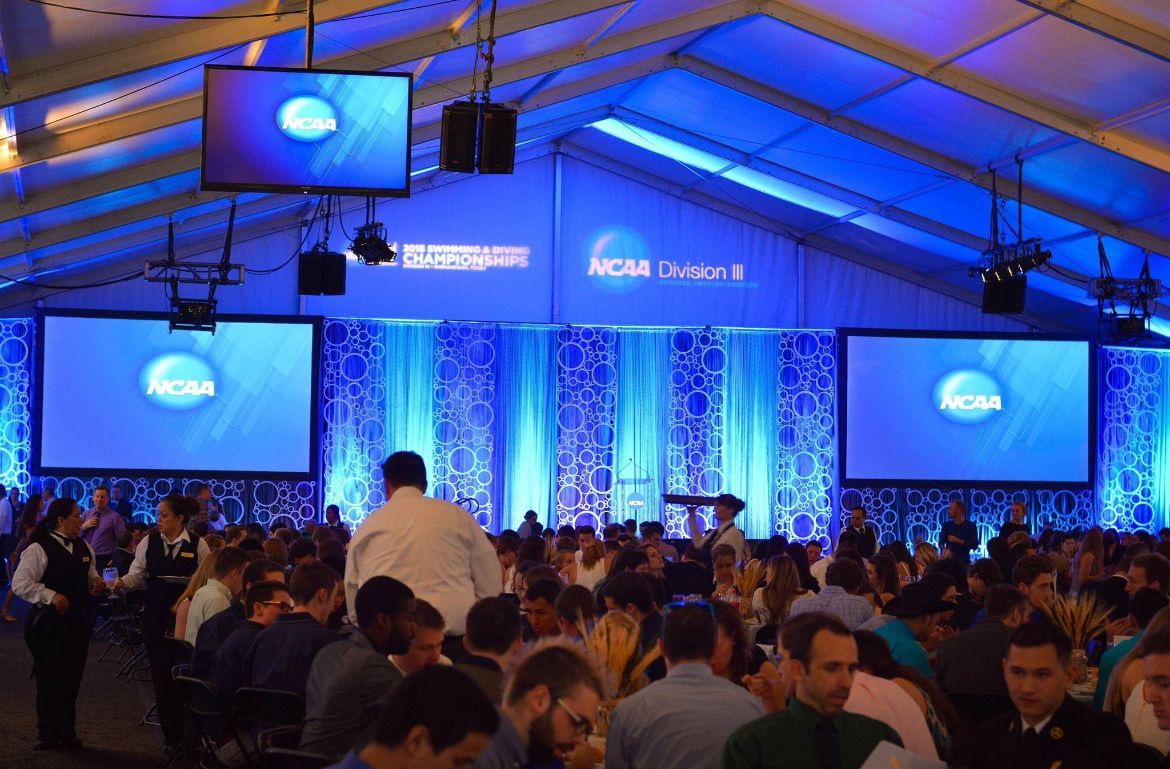 Large crowd at an NCAA Division II event in a tented hall with blue stage lighting and digital screens.