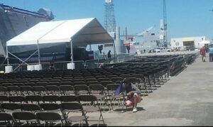 Rows of brown folding chairs set up in front of a white tent stage. Person picking up something.