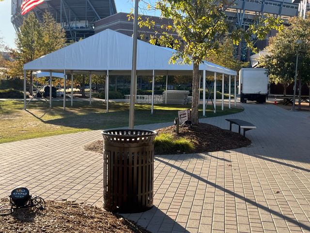 Large white tent set up in a park on a sunny day. A trash can and trees are in the foreground.