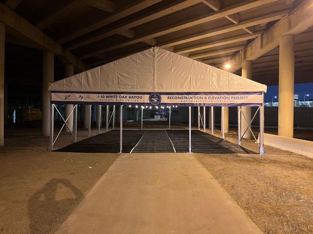 Tent under a bridge, providing shelter. Asphalt walkway leads to it.  Blue and white accents. Evening setting.