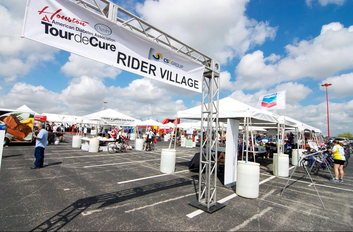 Rider Village at Tour de Cure event with tents, cyclists, and banners under a cloudy sky.