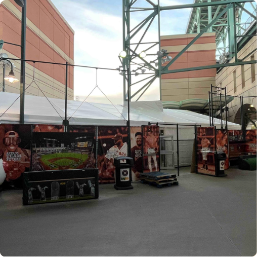 Large posters of baseball players and a stadium view stand before a white event tent at an outdoor ballpark concourse.