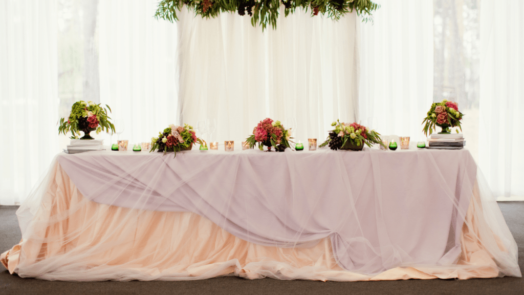 Head table at wedding reception, draped in peach and lavender fabric, adorned with floral arrangements and greenery.