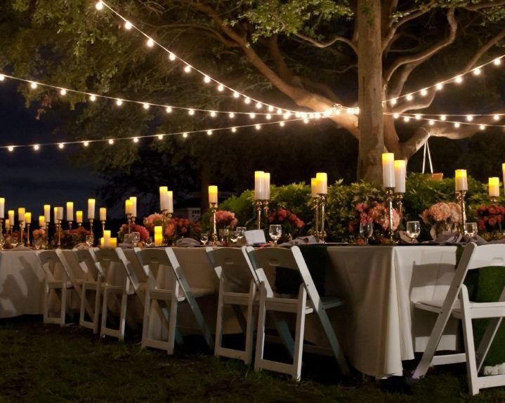 Outdoor dinner party table set with candles and string lights at night.