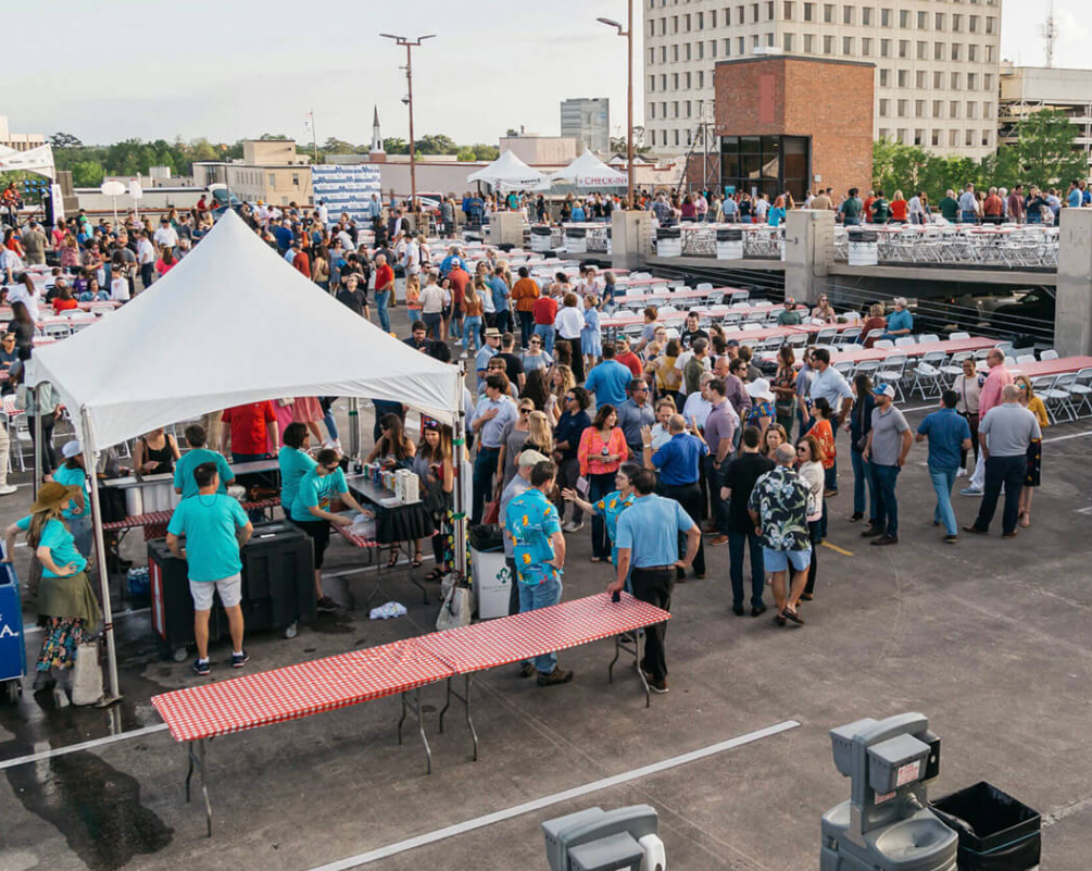 Large crowd at an outdoor event with food tents, picnic tables, and city buildings in the background.