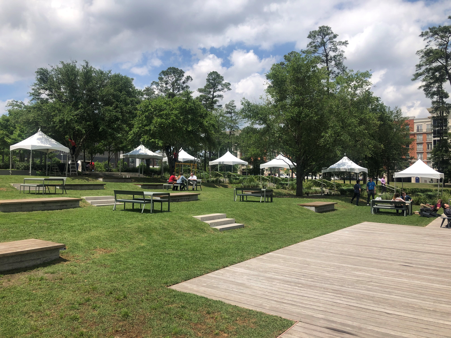 Park scene with white tents, picnic tables, and people enjoying the outdoors on a partly cloudy day.