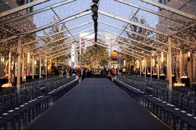 Fashion show runway under a transparent tent. Rows of chairs face a black runway. People gather at the end.