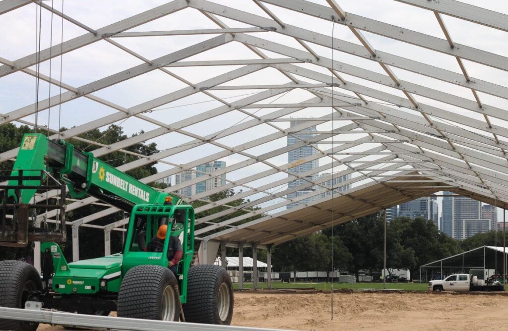 Green forklift assembling a large white tent frame on a brown field. City skyline in the background.