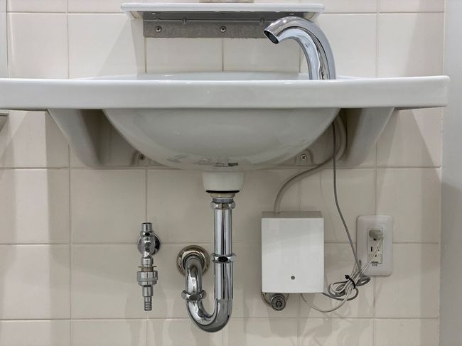 Bathroom sink with exposed plumbing, faucet, soap dispenser, and an electronic device on the wall.