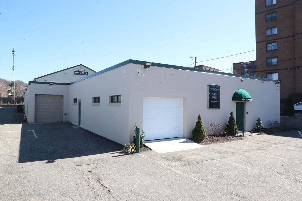 White commercial building with garage doors, green awning, and small shrubs in a parking lot under a clear sky