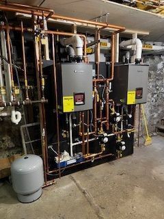 Industrial mechanical room with two gray boilers, exposed copper piping, and a floor tank